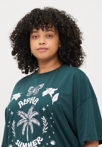 T-shirt oversize color teal con una stampa grafica bianca che presenta una farfalla, dei fiori e una palma, realizzato in tessuto morbido e traspirante.