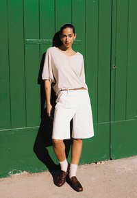 Vrouw met een beige top, witte knielange shorts, bruine loafers en sokken staat in het zonlicht tegen een grote groene houten deur.