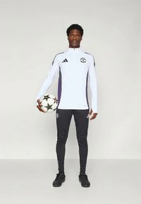 Bijela dugih rukava atletska majica s ljubičastim detaljima, crne trenirke i crne nogometne cipele. Drži crno-bijelu nogometnu loptu.