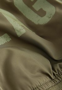 Tissu vert olive à texture lisse avec des lettres vert clair délavées. Détails de fronces au bas.