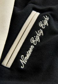 Tejido negro con acentos beige. Presenta texto bordado "Diecinueve Ochenta y Ocho" en una escritura curva. Textura suave, diseño deportivo.