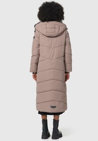 Lange, beige gewatteerde jas met een capuchon. Heeft een chevronstiksel en een merklabel aan de onderrand. Draagt men met zwarte enkellaarsjes.