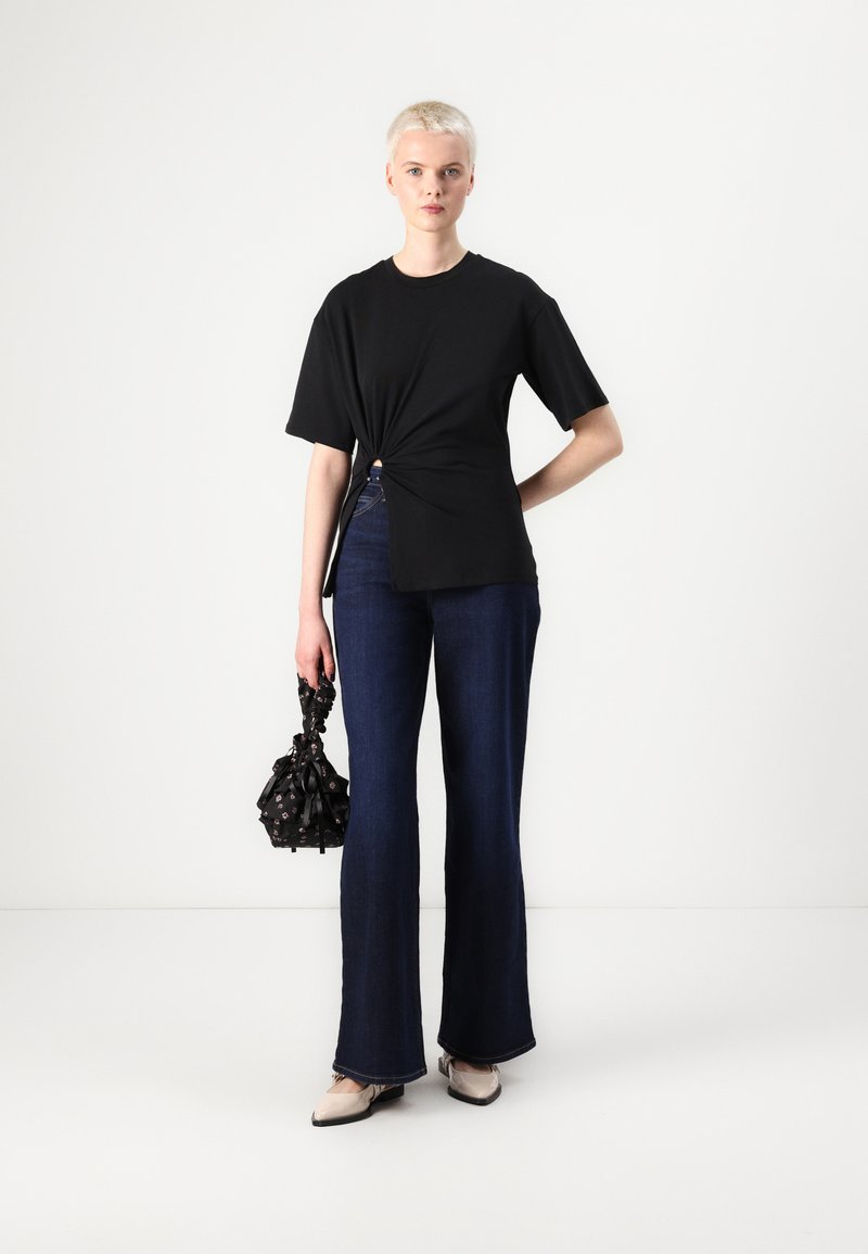 Czarna koszulka z węzłem i krótkim rękawem, w połączeniu z ciemnoniebieskimi dżinsami dzwonami. Akcesorium: mała czarna torba w kwiatowy wzór z wiązaniami.