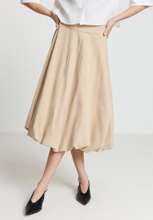 Jupe midi beige à ourlet ballon, portée avec un chemisier blanc, mains sur les hanches, et des talons noirs à bout pointu sur un fond blanc uni.