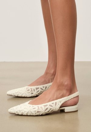 Lábak fehér slingback lapos cipőben, virágos csipke hímzéssel és alacsony blokk sarokkal, sima bézs padlón.