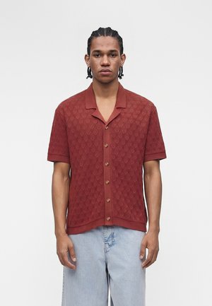 Homme aux cheveux tressés portant une chemise à manches courtes rouille avec un motif texturé et un jean en denim bleu clair.
