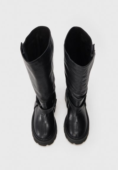 Ψηλές μαύρες δερμάτινες μπότες με χοντρές σόλες και λεπτομέρειες από λουριά, που απεικονίζονται από πάνω σε άσπρο φόντο.