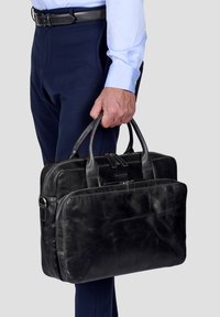 Schwarze Leder-Laptoptasche mit doppelten Griffen, vorderer Reißverschlusstasche, glatter Textur und dezentem Branding. Kombiniert mit marineblauen Hosen und einem hellblauen Hemd.