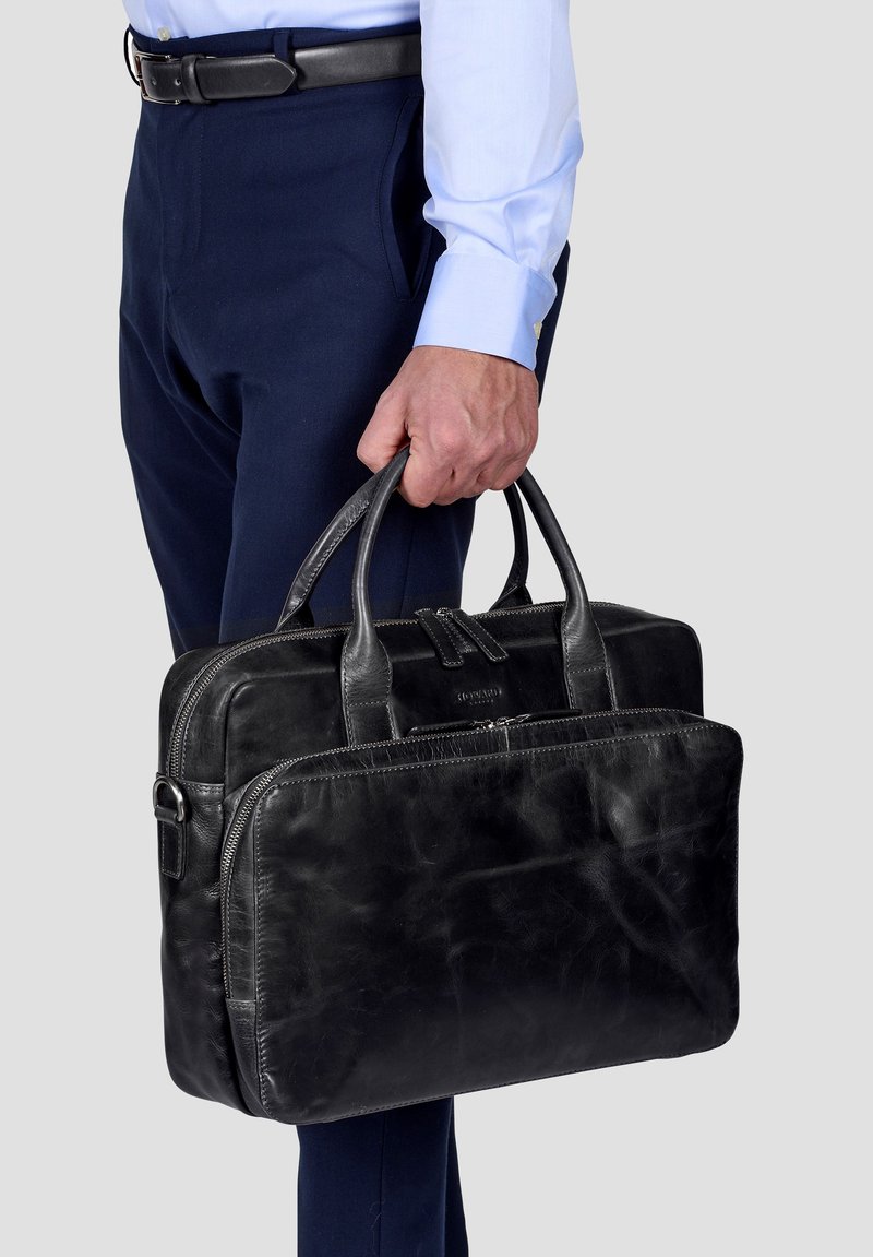 Schwarze Leder-Laptoptasche mit doppelten Griffen, vorderer Reißverschlusstasche, glatter Textur und dezentem Branding. Kombiniert mit marineblauen Hosen und einem hellblauen Hemd.