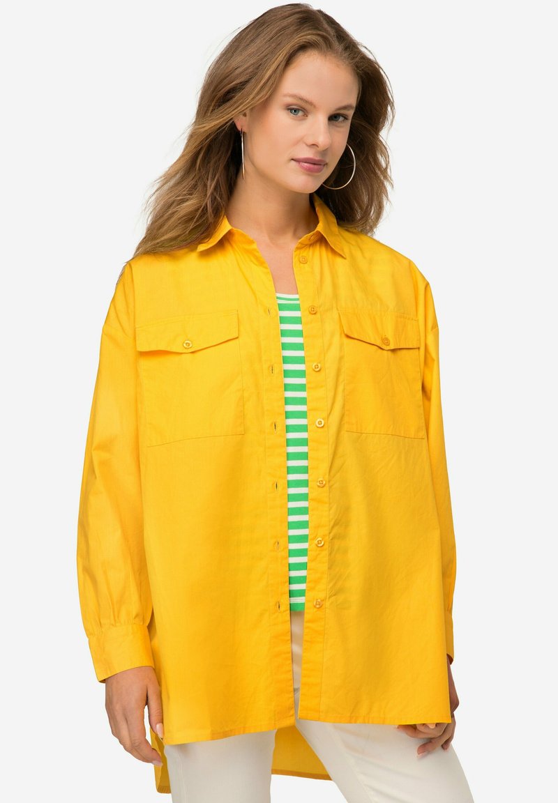Camicia gialla con bottoni, due tasche sul petto e maniche lunghe, realizzata in un tessuto liscio. Indossata sopra una camicia a righe verdi.