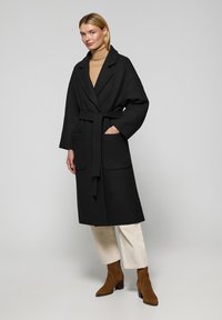 Abrigo negro de manga larga con cintura ceñida, bolsillos laterales y solapa de pico. Combinado con pantalones crema y botines de ante marrón.