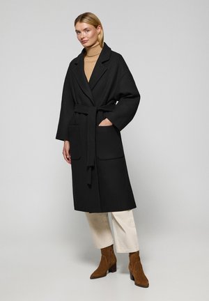 Manteau noir à manches longues avec une taille ceinturée, des poches latérales et un col cranté. Associé à un pantalon crème et des bottes chelsea en daim marron.