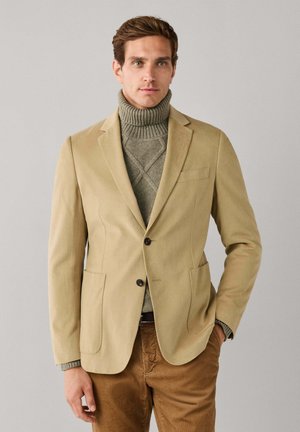 Mann trägt einen beigefarbenen Blazer über einem grauen Rollkragenpullover und braune Hosen, steht mit einer Hand in der Tasche vor einfarbigem Hintergrund.