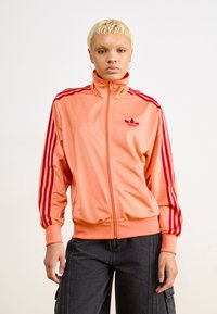 Orange Zip-Jacke aus glattem Material, mit roten Streifen an den Schultern und Ärmeln, sowie einem kleinen roten Logo auf der Brust.