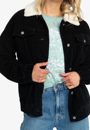 Kobieta w czarnej sztruksowej kurtce z białym kołnierzem z polaru na wzorzystej miętowej koszuli i niebieskich dżinsach, trzymająca krawędzie kurtki.