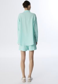 Camisa verde menta texturizada con cuello y corte holgado, combinada con pantalones cortos a juego. Calzado casual visible. Diseño simple, con detalles mínimos.