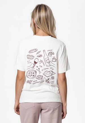 Lyshåret kvinde iført en hvid t-shirt med håndtegnede illustrationer af fransk mad og drikke på ryggen.