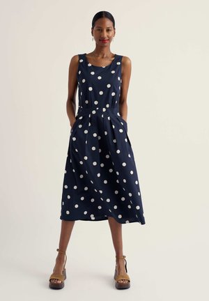 Mujer de pie con las manos en los bolsillos, que lleva un vestido midi sin mangas azul marino con lunares blancos y sandalias de plataforma color camel.