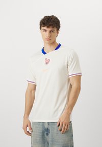 Biała koszulka sportowa z niebieskim kołnierzykiem i czerwono-niebieskimi paskami na rękawach. Na przodzie znajduje się logo koguta oraz emblemat Nike.