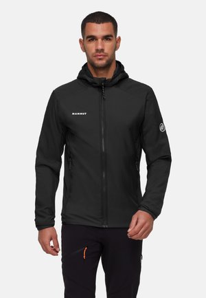 Man wearing a black Mammut hooded jacket with front zipper, standing against a plain light background.