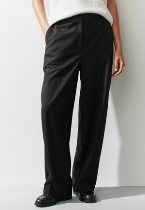 Kvinde iført sorte skræddersyede vide bukser, hvid sweater og sorte loafers, stående mod en ensfarvet hvid væg.