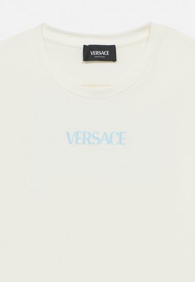 Balta T-krekls ar apaļu izgriezumu, priekšpusē centrā gaiši zilā krāsā redzams "VERSACE" logotips un melna etiķete ar uzrakstu "VERSACE Made in Italy" apkakles iekšpusē.