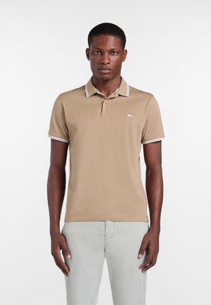 Uomo che indossa una polo beige con bordo bianco e pantaloni grigio chiaro, in piedi davanti a uno sfondo bianco uniforme.