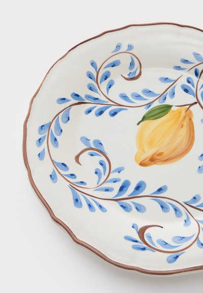 Assiette en céramique blanche avec un bord festonné marron, ornée de volutes feuillues bleues et d’un fruit jaune central avec une feuille verte.