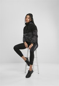 Svart fleece- och glänsande nylonpulloverjacka med hög krage, kombinerad med svarta leggings som har sidotext och svarta sneakers.