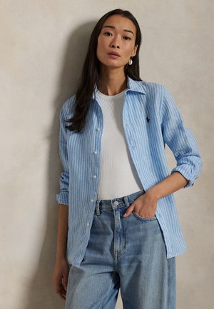 Kvinne med langt mørkt hår har på seg en lys blå, stripete skjorte med knapper over en hvit topp og høytlivede blå jeans, og står mot en beige vegg.