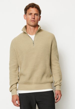 Beige gerippter Pullover mit Stehkragen und halbem Reißverschluss, langen Ärmeln und einem weichen, strukturierten Stoff. Über einem weißen Hemd getragen.