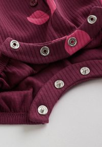 Gros plan sur un tissu côtelé bordeaux avec des empreintes de lèvres roses et des boutons-pression en métal partiellement attachés sur le bord d'un vêtement.