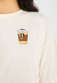 Krämfärgad t-shirt med en cartoon-design av en kaffekopp med ett leende ansikte, svarta ögon och ett brunt papperssleeve.