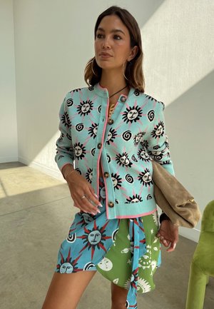 Nő türkiz kardigánban és vegyes mintás szoknyában, amely nap- és égi motívumokat ábrázol, bézs borítéktáskát tart egy napfényes szobában.