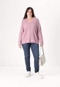 Light pink V-neck sweater with cable knit design, paired with dark blue jeans and white sneakers, holding a light gray handbag.