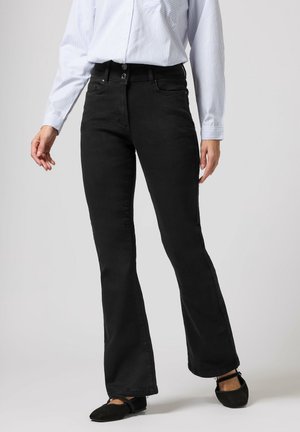 Kobieta w czarnych spodniach bootcut z wysokim stanem, białej koszuli w paski z długim rękawem oraz czarnych balerinach na jasnoszarym tle.