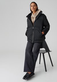 Giacca a vento nera con colletto in peluche, chiusura con zip e due tasche frontali, indossata sopra un maglione beige e abbinata a pantaloni jeans larghi scuri.