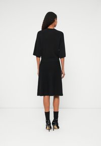 Fekete kötött ruha könyök hosszúságú ujjal, derékpánttal és bővülő szegéllyel. Fekete magassarkú cipővel és bokáig érő zoknival viselve.