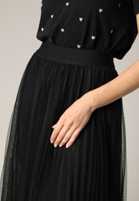 Jupe en tulle noir avec une taille haute élastique et une texture superposée et transparente, avec une main reposant sur la surface du tissu.