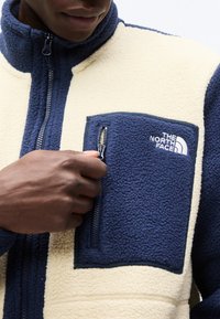 Άτομο που φοράει μπεζ και ναυτικό fleece μπουφάν, τραβώντας το φερμουάρ της τσέπης στο στήθος με το λογότυπο "The North Face".