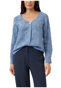 Lichtblauwe, gestructureerde cardigan met een V-hals, lange mouwen en een enkele knoopsluiting, gedragen over een donkere broek.