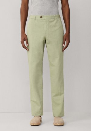 Persona vistiendo pantalones rectos verde claro, camisa gris metida por dentro y alpargatas beige, de pie contra un fondo liso.