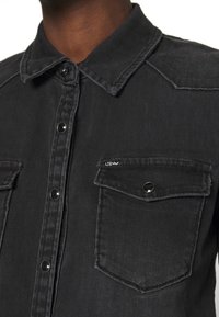 Primo piano di una camicia di denim nera con bottoni a pressione in metallo e tasche a pattina sul petto, indossata da una persona con pelle di colore marrone medio.