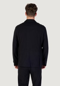 Uomo con capelli ricci che indossa una giacca nera e pantaloni abbinati, in piedi con la schiena rivolta verso uno sfondo chiaro e uniforme.