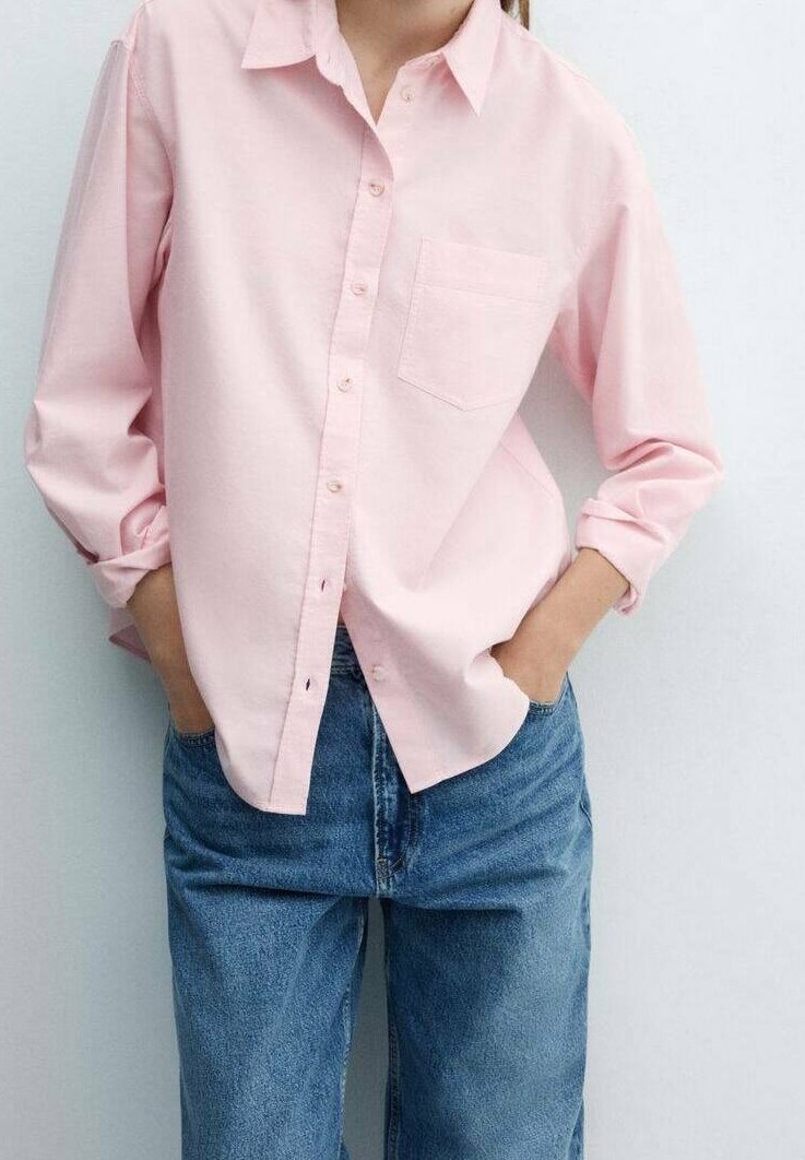 Personne portant une chemise rose pâle à boutons avec les manches retroussées et un jean en denim bleu, les mains dans les poches avant.