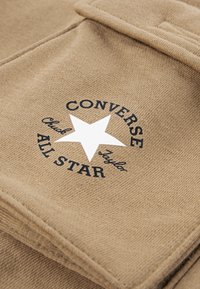 Beige stoff med en rund logo med en hvit stjerne og svart tekst som lyder "CONVERSE Chuck Taylor ALL STAR." Teksturert overflatedetalj synlig.