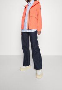 Heller korallfarbener Mantel mit Kapuze, Reißverschluss vorne und zwei Taschen, kombiniert mit einem hellblauen gestreiften Hemd und dunklen, weiten Jeans. Cremefarbene Plateausneaker.