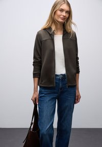 Tmavě zelená mikina na zip s kapucí, nošená přes bílou košili, doplněná modrými širokými džíny. Modelka drží hnědou tašku.
