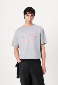 Grå bomull T-skjorte med korte ermer, med rød brodert tekst "TOMMY JEANS NEW YORK CITY" på fronten. Løs passform.