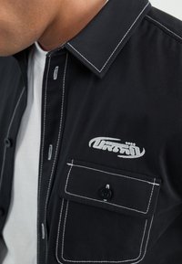 Černá košile s bílým prošíváním, zapínání na knoflíky a náprsní kapsa s vyšitým logem. Bavlněný materiál, strukturovaný design límce.